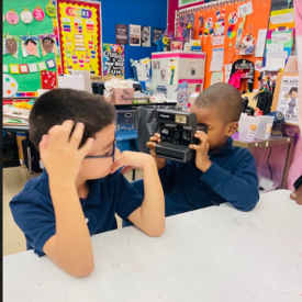 In a grade 1 art classroom, a male student positions a Polaroid camera to his eye as he takes a photo of a male classmate seated at a table.